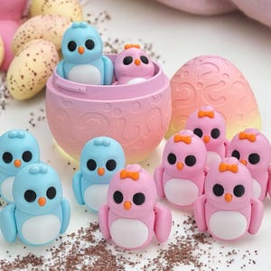 May include: A set of pastel blue and pink bird figurines with bows. The figurines are arranged around a pink and orange egg-shaped container with a lid. The container is decorated with a swirl pattern.