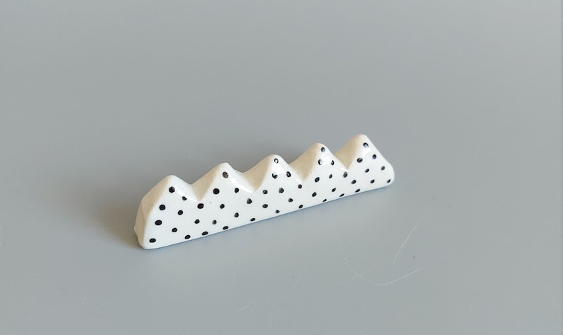 May include: White ceramic chopstick rest with a black polka dot pattern shaped like a row of mountains.