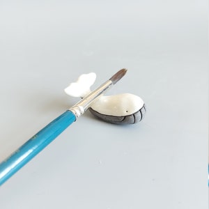 May include: A small white ceramic whale figurine with a gray belly, resting on a blue paintbrush.