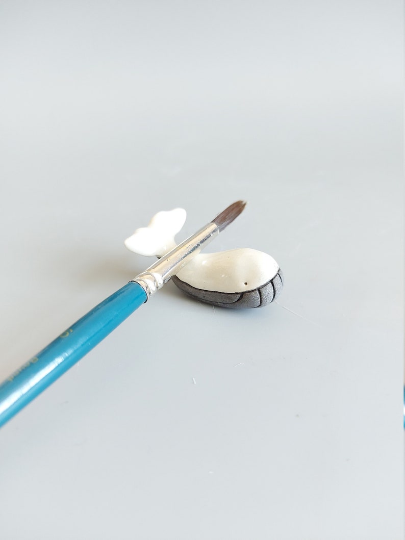 May include: A small white ceramic whale figurine with a gray belly, resting on a blue paintbrush.