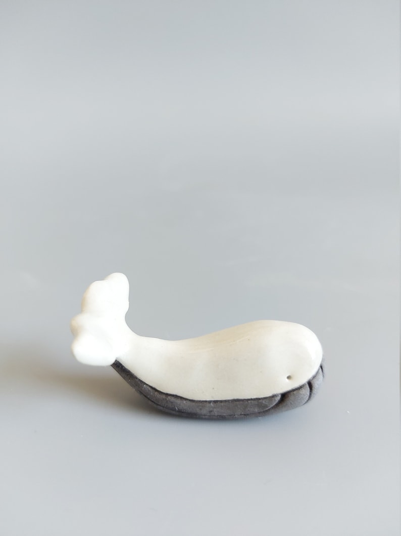 May include: A white ceramic whale figurine with a gray underside. The whale is lying on its side with its tail in the air.