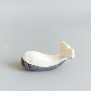 May include: A small white ceramic whale figurine with a gray underside. The whale has a simple, stylized design and a small tail fin.