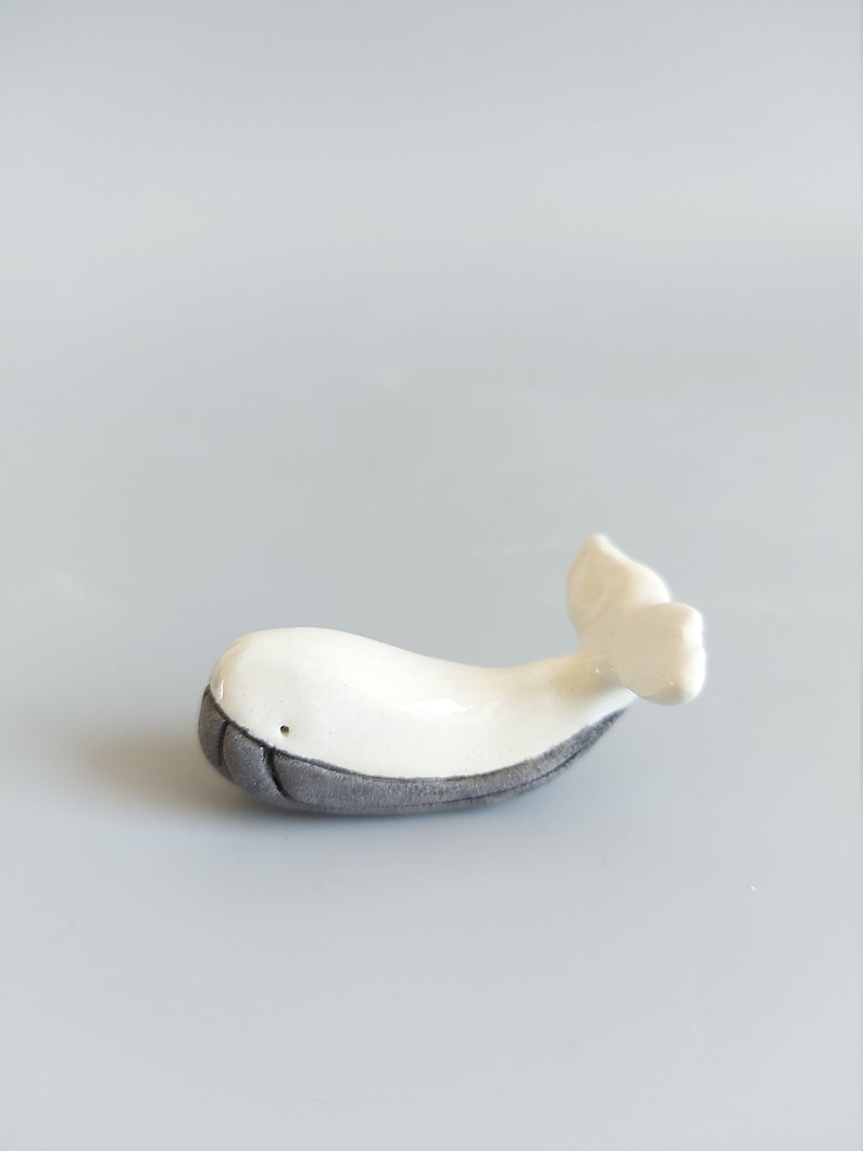 May include: A small white ceramic whale figurine with a gray underside. The whale has a simple, stylized design and a small tail fin.