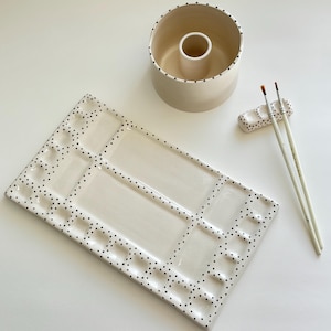 Puede incluir: Un juego de artista de cerámica con una paleta rectangular con pozos, un recipiente cilíndrico para agua, un soporte para pinceles y dos pinceles. El juego es de color blanco roto con lunares negros, ideal para pintar.