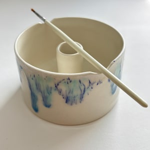 Puede incluir: Un soporte de cerámica para pinceles con un compartimento cilíndrico central y una cuenca circular circundante. El soporte blanquecino tiene goteos azules y turquesas alrededor del borde. Un pincel descansa en la parte superior.