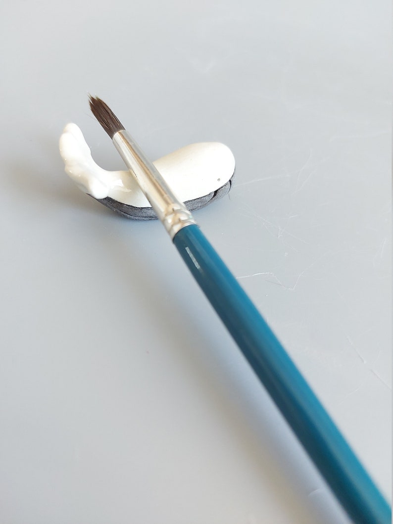 May include: A white ceramic whale brush holder with a blue paintbrush resting on top.
