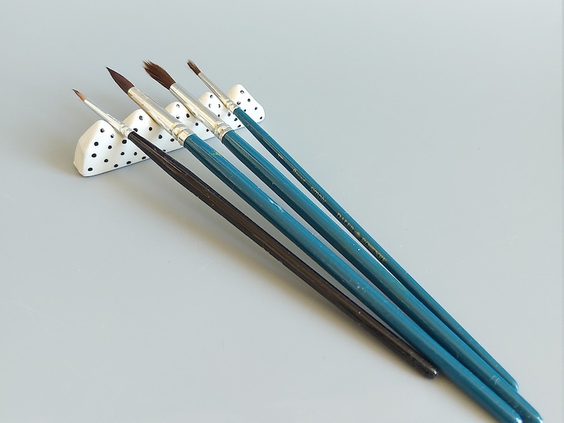 May include: Five paintbrushes in a white ceramic holder with black polka dots. The brushes have teal blue handles and brown bristles.