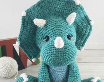 Op de afbeelding: Een turquoise amigurumi dinosaurus met witte accenten, waaronder hoorns en voetdetails. De gehaakte dinosaurus heeft een grote kraag en zit rechtop. De tekst "Dinosaur Amigurumi Pattern" staat bovenaan de afbeelding.