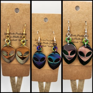 May include: Three pairs of alien-shaped earrings with iridescent finishes. The earrings are made of metal and have a black backing. The earrings are hanging from silver hooks.