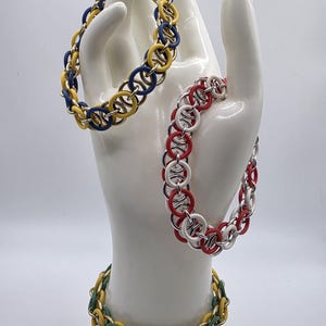 May include: Three colorful chainmail bracelets. One bracelet features yellow and blue rings, another has red and white rings, and the third has green and yellow rings. The bracelets are displayed on a white hand-shaped jewelry holder.