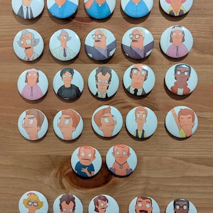 May include: A collection of 27 round button pins featuring various cartoon characters. The characters are all male and have different hairstyles, facial expressions, and clothing. The buttons are white with a black outline and are arranged in a grid pattern on a wooden surface.