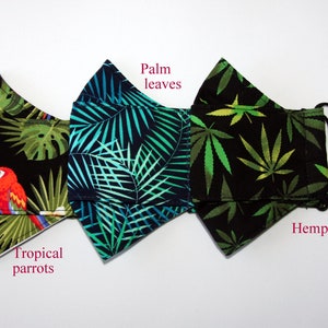 Puede incluir: Tres mascarillas negras con diferentes estampados tropicales. La mascarilla de la izquierda tiene un dise&ntilde;o de loro tropical, la del medio tiene hojas de palma y la de la derecha tiene hojas de c&aacute;&ntilde;amo. Las mascarillas tienen lazos el&aacute;sticos negros para las orejas.