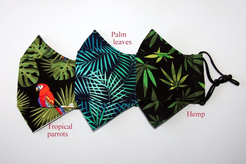 Puede incluir: Tres mascarillas negras con diferentes estampados tropicales. La mascarilla de la izquierda tiene un dise&ntilde;o de loro tropical, la del medio tiene hojas de palma y la de la derecha tiene hojas de c&aacute;&ntilde;amo. Las mascarillas tienen lazos el&aacute;sticos negros para las orejas.