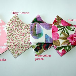 Puede incluir: Cuatro mascarillas de tela diferentes con patrones florales. Las mascarillas son de color rosa, blanco y verde con varios dise&ntilde;os florales, incluyendo margaritas, lirios y un patr&oacute;n de jard&iacute;n de acuarela. Las mascarillas est&aacute;n etiquetadas con sus nombres: Smiling daisies, Ditsy flowers, Watercolor garden y Pink lilies.