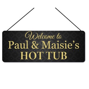 May include: Black rectangular sign with gold text that reads "Welcome to Paul & Maisie's HOT TUB." The sign has a diamond pattern background and is suspended by two black strings.