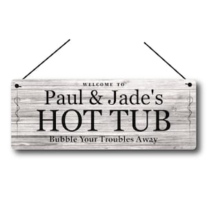 Könnte beinhalten: Rechteckiges Holzschild mit dem Text "Welcome to Paul & Jade's HOT TUB Bubble Your Troubles Away" in schwarzer Schrift. Das Schild hat eine verwitterte, graue Holzmaserung und ist an schwarzen Schnüren aufgehängt.