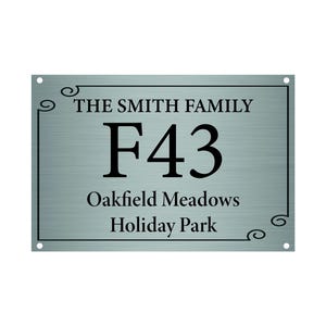 May include: A rectangular metal sign with the text "THE SMITH FAMILY", "F43", "Oakfield Meadows", and "Holiday Park" in black. The sign has a brushed metal appearance with decorative swirls and four mounting holes.