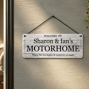 May include: A rectangular wooden sign with the text "WELCOME TO Sharon & Ian's MOTORHOME" in black lettering. Below, it reads "Where the fun begins & memories are made." The sign is hanging on a wall.