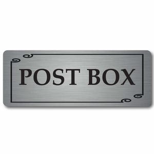 May include: A rectangular, brushed silver sign with the words "POST BOX" in bold, black capital letters. The sign has a black border with decorative swirls in the corners. The background is white.