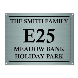 May include: A rectangular metal sign with the text "THE SMITH FAMILY", "E25", "MEADOW BANK", and "HOLIDAY PARK" in black font. The sign has a brushed metal appearance and is mounted with four corner holes.