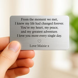 May include: A rectangular, brushed metal card with engraved text. The text reads, "From the moment we met...I love you more every single day." A heart symbol and the name "Maisie" are also included. The card is held by a person.