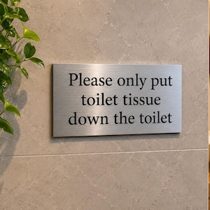 May include: A rectangular, brushed metal sign with the text "Please only put toilet tissue down the toilet" in black lettering. The sign is mounted on a light grey tiled wall. A green plant is visible on the left.
