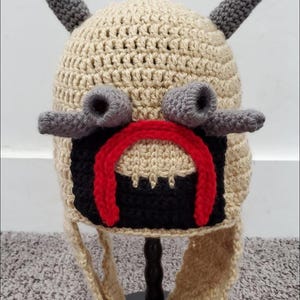May include: A handmade crochet hat in tan, gray, black, and red. The hat features two gray horns, gray nose-like protrusions, and a red and black face design. The hat has ear flaps and is displayed on a black stand.