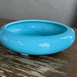 May include: A shallow, round ceramic bowl with a vibrant turquoise blue glossy glaze. The bowl features a wide, smooth rim and a low-profile design, resting on small, subtle feet that elevate the base slightly from the surface.