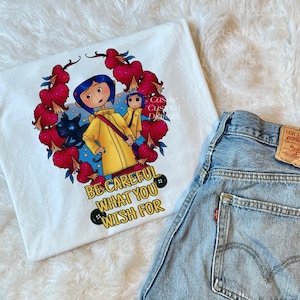 May include: White t-shirt with a colorful illustration of a girl in a yellow raincoat, a black cat, and a small monkey. The illustration is surrounded by red flowers and the text "Be careful what you wish for".