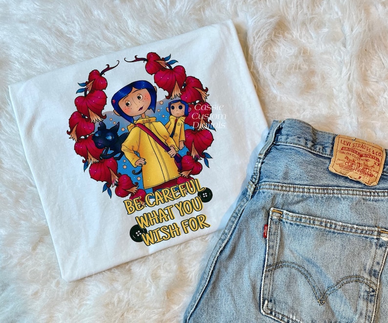 May include: White t-shirt with a colorful illustration of a girl in a yellow raincoat, a black cat, and a small monkey. The illustration is surrounded by red flowers and the text "Be careful what you wish for".