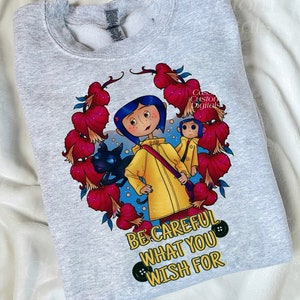 May include: A gray sweatshirt with a colorful illustration of a girl in a yellow raincoat with a black cat and a monkey. The text "Be careful what you wish for" is printed below the illustration.