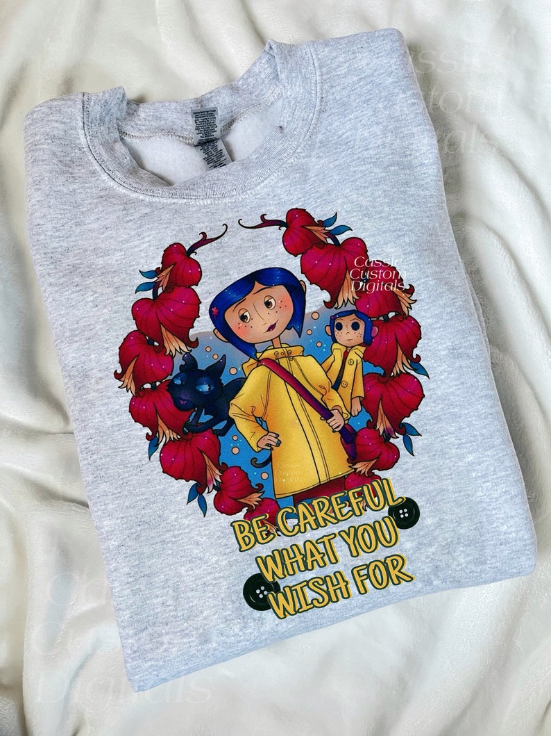 May include: A gray sweatshirt with a colorful illustration of a girl in a yellow raincoat with a black cat and a monkey. The text "Be careful what you wish for" is printed below the illustration.