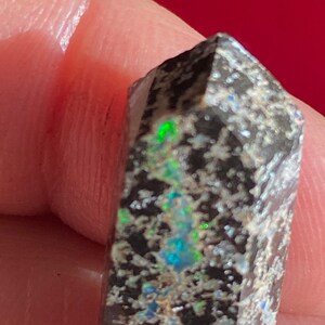 May include: A dark, faceted gemstone with a rough, speckled surface. The stone displays flashes of green, blue, and iridescent colors. The gemstone is a small, polished crystal, likely used for jewelry or collecting.