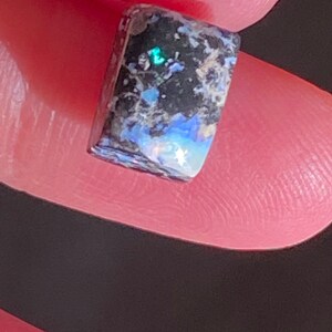 May include: A rectangular black opal gemstone with iridescent blue, green, and white flashes. The stone has a dark base with lighter, speckled inclusions. The opal is held between fingers, showcasing its polished surface and natural beauty.