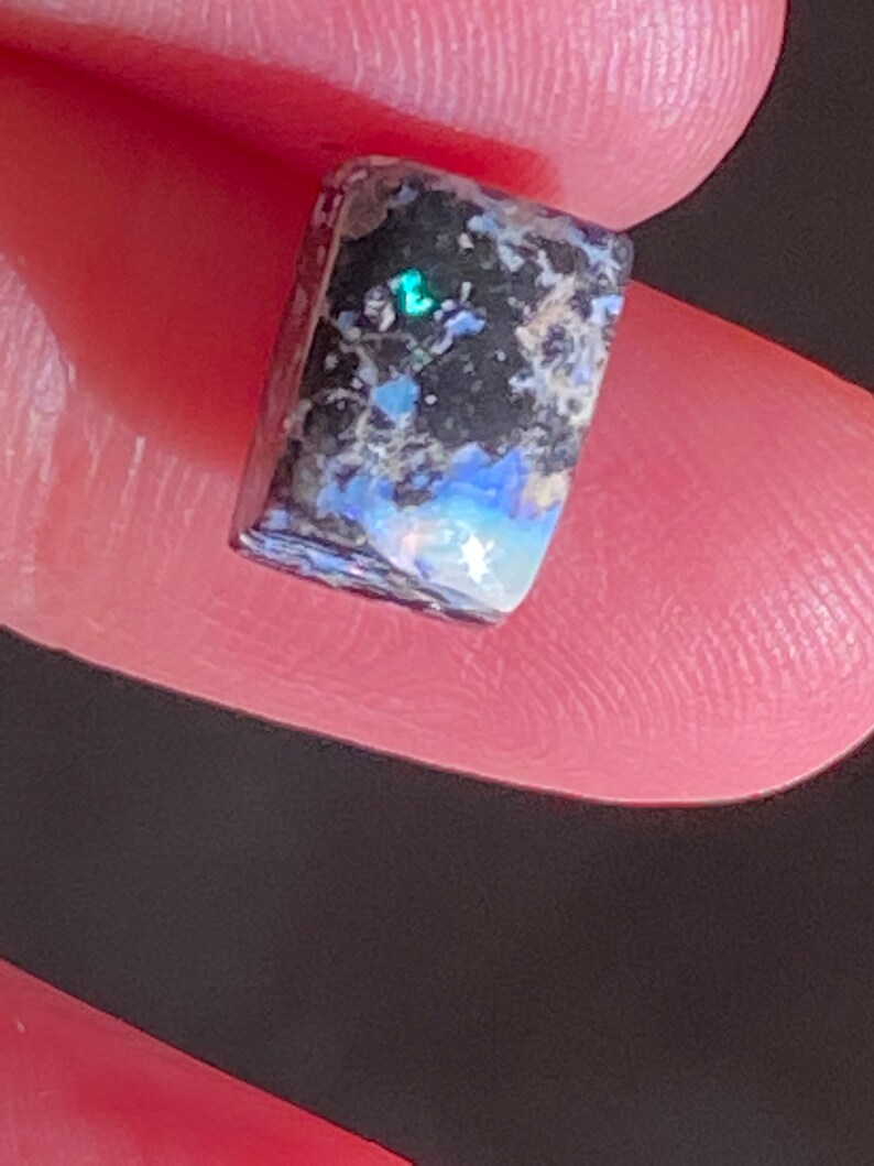 May include: A rectangular black opal gemstone with iridescent blue, green, and white flashes. The stone has a dark base with lighter, speckled inclusions. The opal is held between fingers, showcasing its polished surface and natural beauty.