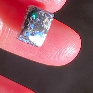May include: A small, rectangular gemstone with a dark, speckled surface. The stone has a vibrant green flash in the center, with hints of blue and white throughout. The gemstone is held between two fingers.