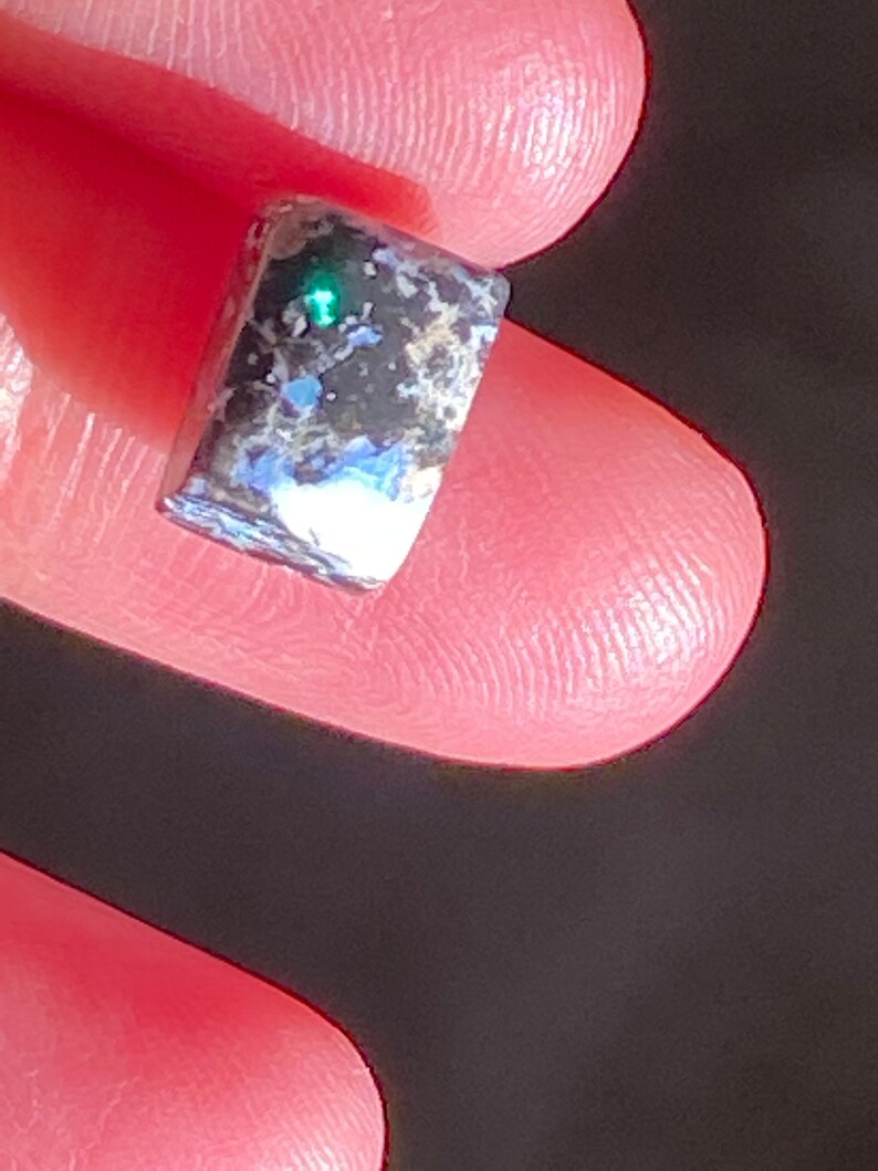 May include: A small, rectangular gemstone with a dark, speckled surface. The stone has a vibrant green flash in the center, with hints of blue and white throughout. The gemstone is held between two fingers.