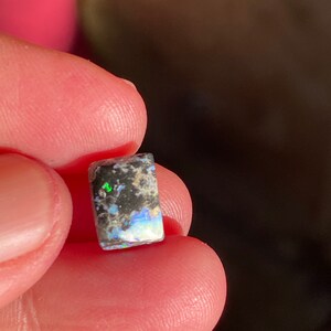 May include: A small, rectangular opal gemstone with a mix of black, white, and iridescent colors. The opal is held between fingers, showcasing its polished surface and natural patterns. The gemstone displays flashes of green and blue.