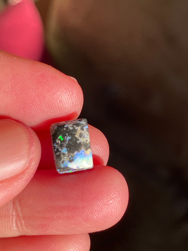 May include: A small, rectangular opal gemstone with a mix of black, white, and iridescent colors. The opal is held between fingers, showcasing its polished surface and natural patterns. The gemstone displays flashes of green and blue.