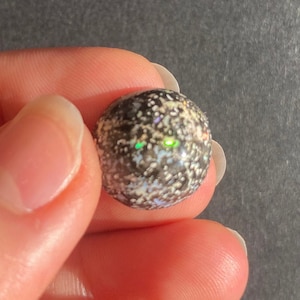 May include: A round, dark-colored gemstone with a speckled pattern and iridescent highlights. The stone is held between fingers against a gray background, showcasing its texture and color variations. The gem's surface reflects a vibrant green hue.