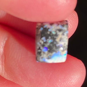 May include: A rectangular opal gemstone with a dark base color, speckled with iridescent blues, greens, and whites. The stone is held between two fingers, showcasing its polished surface and unique color play.