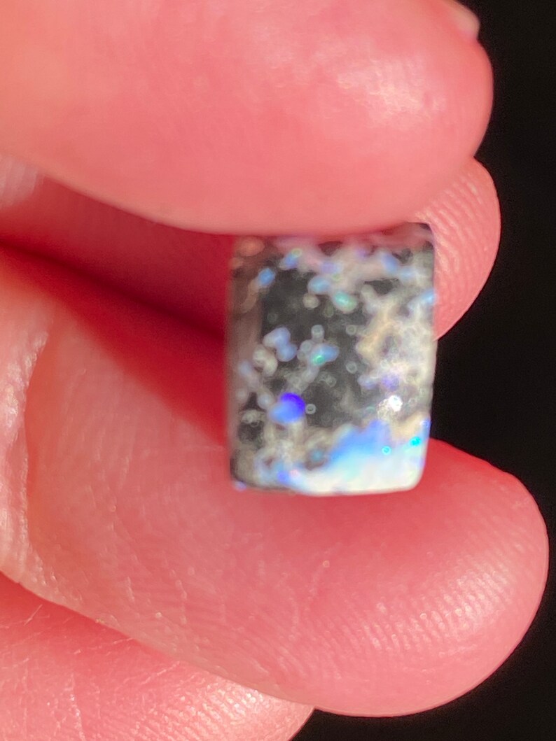May include: A rectangular opal gemstone with a dark base color, speckled with iridescent blues, greens, and whites. The stone is held between two fingers, showcasing its polished surface and unique color play.