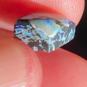 May include: A small, faceted gemstone with a dark gray base and iridescent blue, green, and white patterns. The stone is held between fingers, showcasing its unique color play and texture. The gem's cut reveals multiple facets.