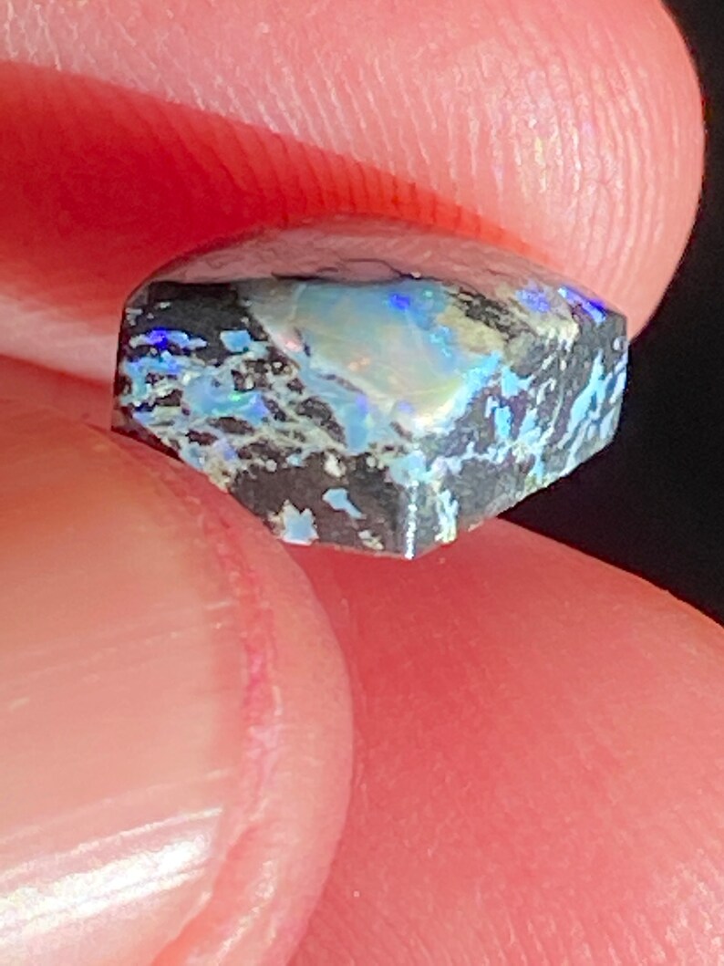 May include: A small, faceted gemstone with a dark gray base and iridescent blue, green, and white patterns. The stone is held between fingers, showcasing its unique color play and texture. The gem's cut reveals multiple facets.