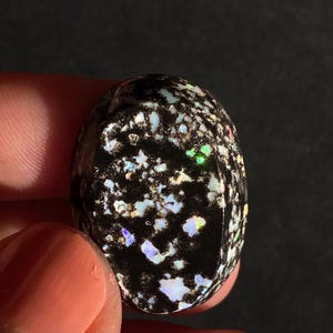 May include: An oval-shaped gemstone with a black base and a speckled pattern of white, blue, and green iridescent inclusions. The polished surface reflects light, creating a shimmering effect. The gemstone is held between fingers.