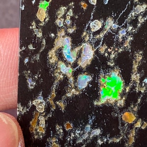 May include: Close-up of a black stone with a unique pattern. The stone features a dark background with scattered, colorful inclusions of green, blue, and orange. The inclusions have a shimmering, iridescent quality, creating a striking visual effect. The stone's surface is slightly textured.