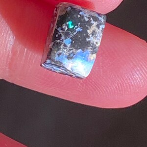 May include: A rectangular gemstone with a polished surface. The stone features a mix of dark gray, black, and blue hues, with iridescent flashes of green and blue. The gemstone is held between two fingers.