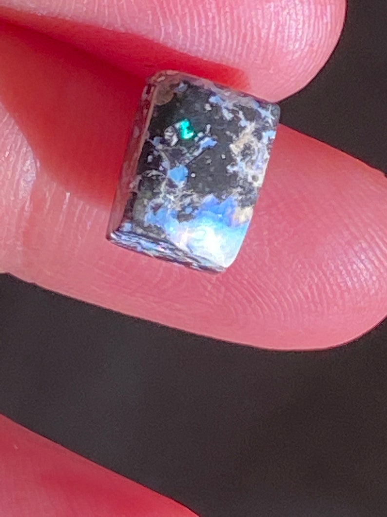 May include: A rectangular gemstone with a polished surface. The stone features a mix of dark gray, black, and blue hues, with iridescent flashes of green and blue. The gemstone is held between two fingers.
