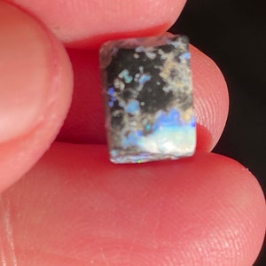May include: A small, rectangular opal gemstone with a black base and iridescent blue, green, and white inclusions. The opal is held between two fingers, showcasing its natural colors and textures.