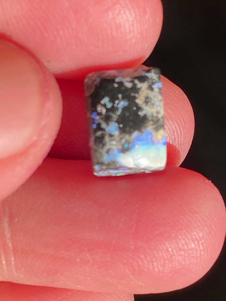 May include: A small, rectangular opal gemstone with a black base and iridescent blue, green, and white inclusions. The opal is held between two fingers, showcasing its natural colors and textures.